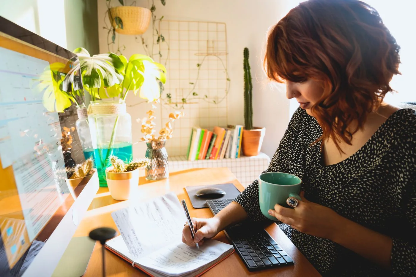 Woman writing in a notebook while holding coffee at a home desk