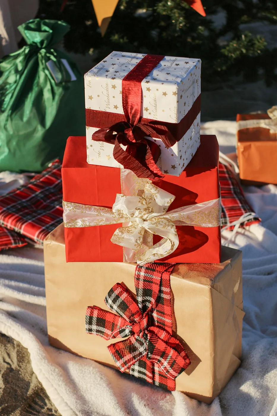 Stacked wrapped gifts with red and pale ribbons beside a decorated tree.