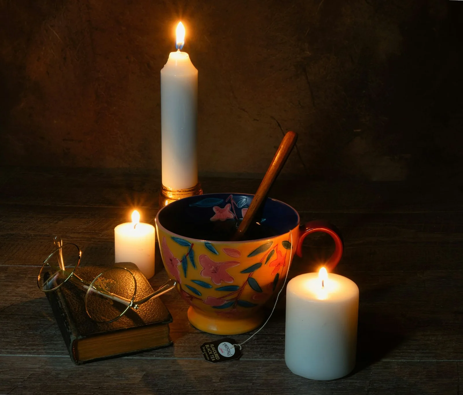 A colourful cup beside a small book, glasses and lit candles.