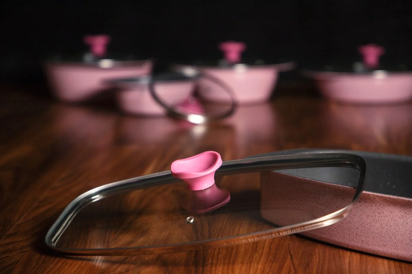 Pink cooking pots and pans with glass lids on a wooden surface.
