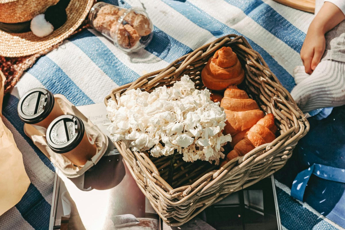 Picnic food and drinks beside a wicker basket on a striped blanket.