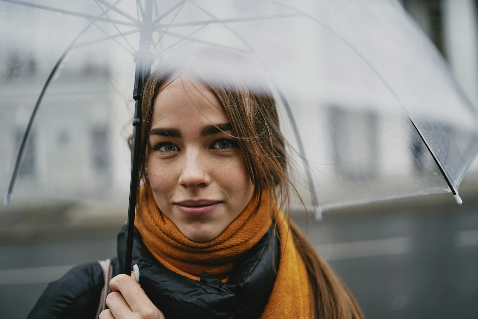 A woman wearing a scarf beneath a clear umbrella.